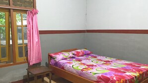 Une petite chambre avec un lit simple, un cadre en bois, une couvre-lit à motifs floraux colorés, un petit tabouret en bois et une fenêtre ornée d’un rideau rose.