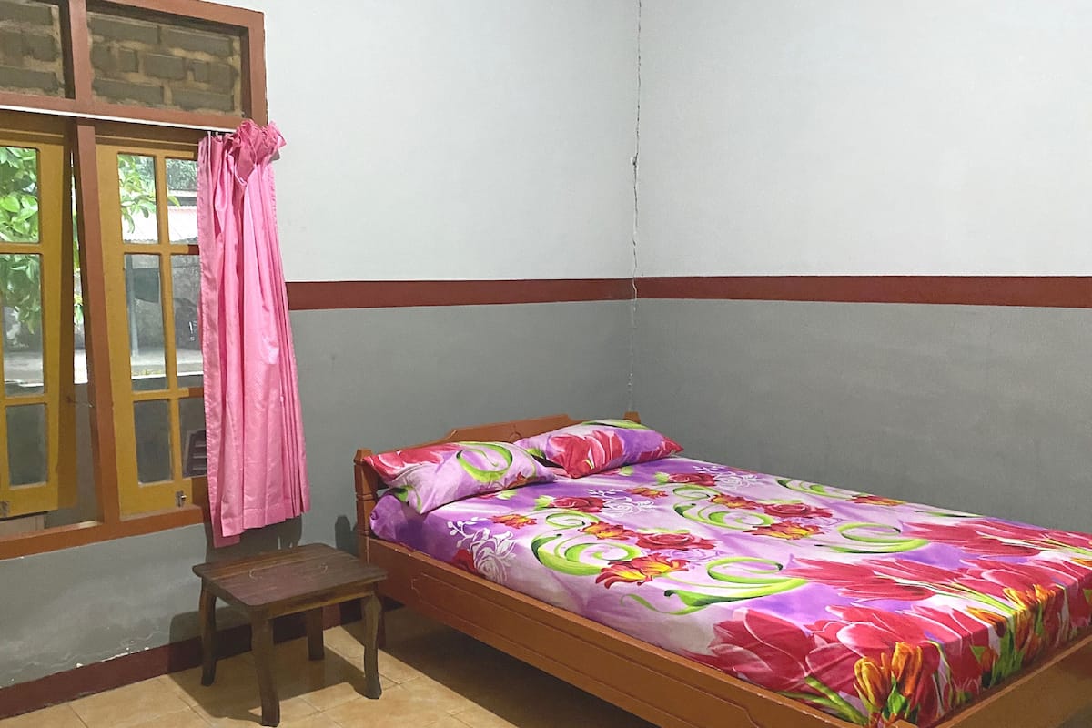 Une petite chambre avec un lit simple, un cadre en bois, une couvre-lit à motifs floraux colorés, un petit tabouret en bois et une fenêtre ornée d’un rideau rose.