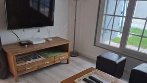 Ein Wohnzimmer mit einem Holz-Fernsehschrank, einem Flachbildfernseher, einer Stehlampe und einem Fenster mit Blick ins Freie.