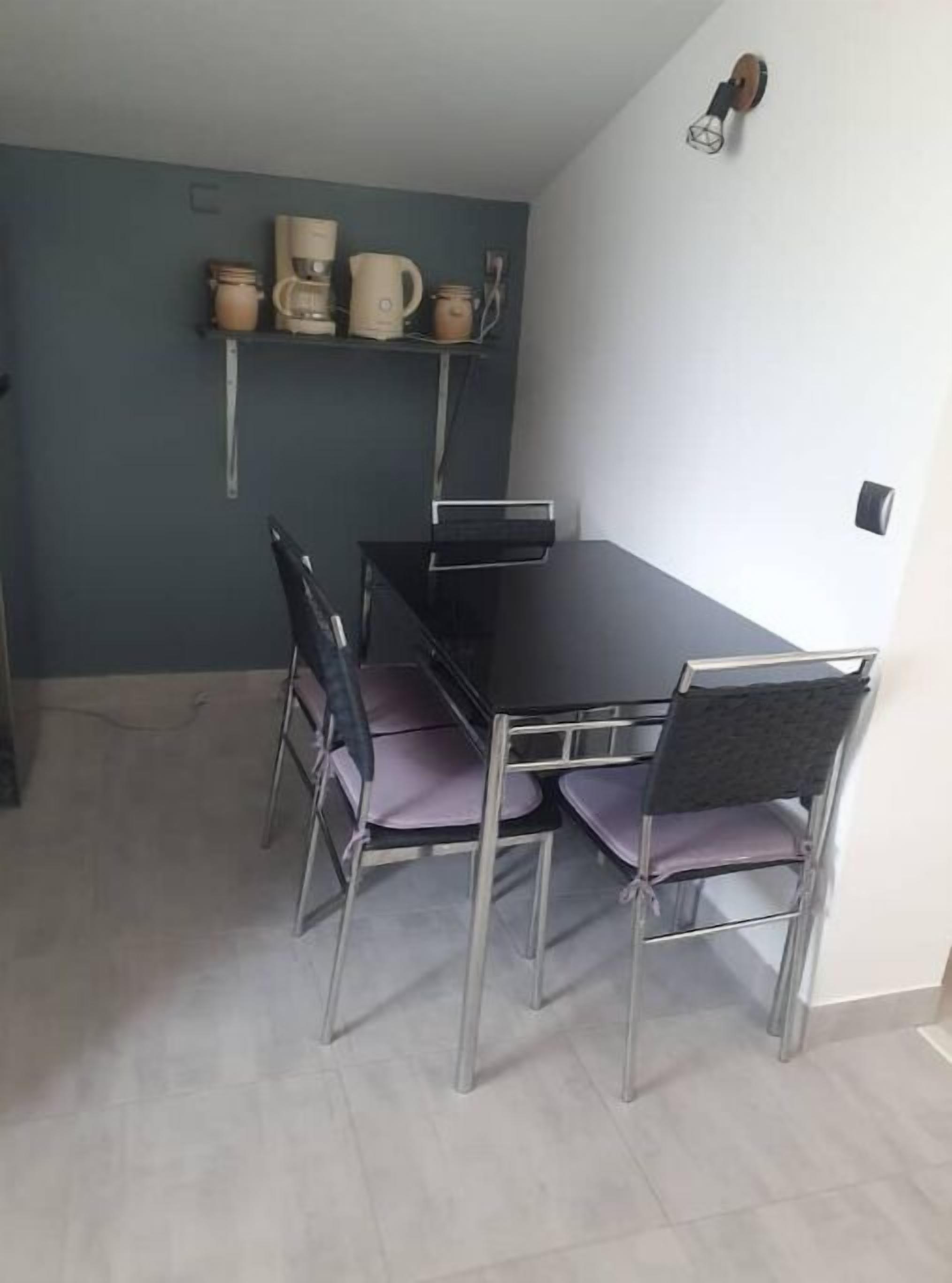 A dining area with a black table and four chairs, a shelf with kitchen items, and a light switch on the wall.