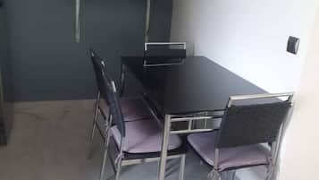 A dining area with a black table and four chairs, a shelf with kitchen items, and a light switch on the wall.