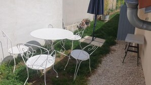 Eine Terrasse mit einem weißen Metalltisch und -stühlen, einem blauen Sonnenschirm und einer kleinen Gartenanlage mit Blumentöpfen.