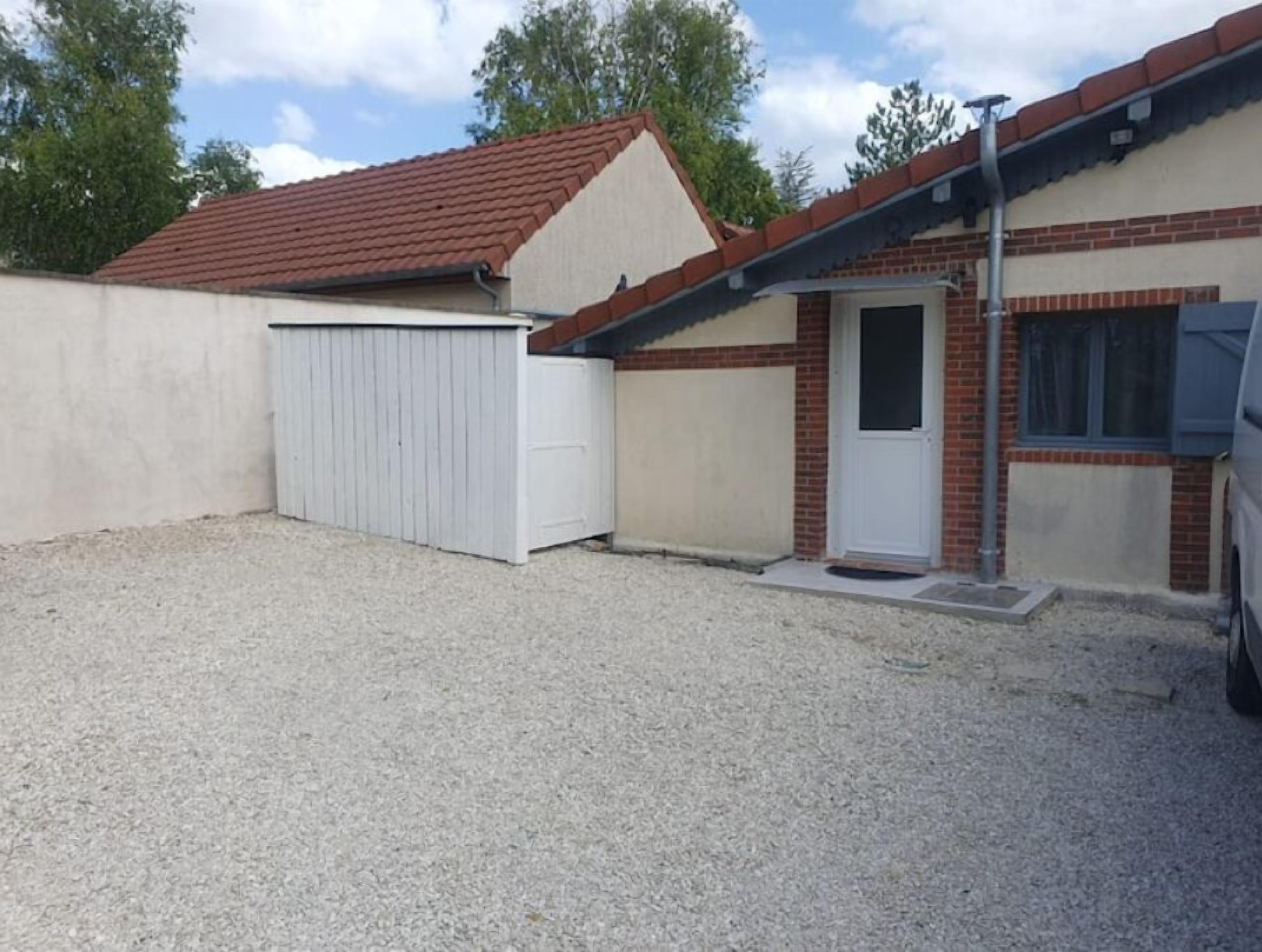 Un bâtiment d’un seul étage, avec un toit recouvert de tuiles rouges, une porte de garage blanche et une petite porte d’entrée.