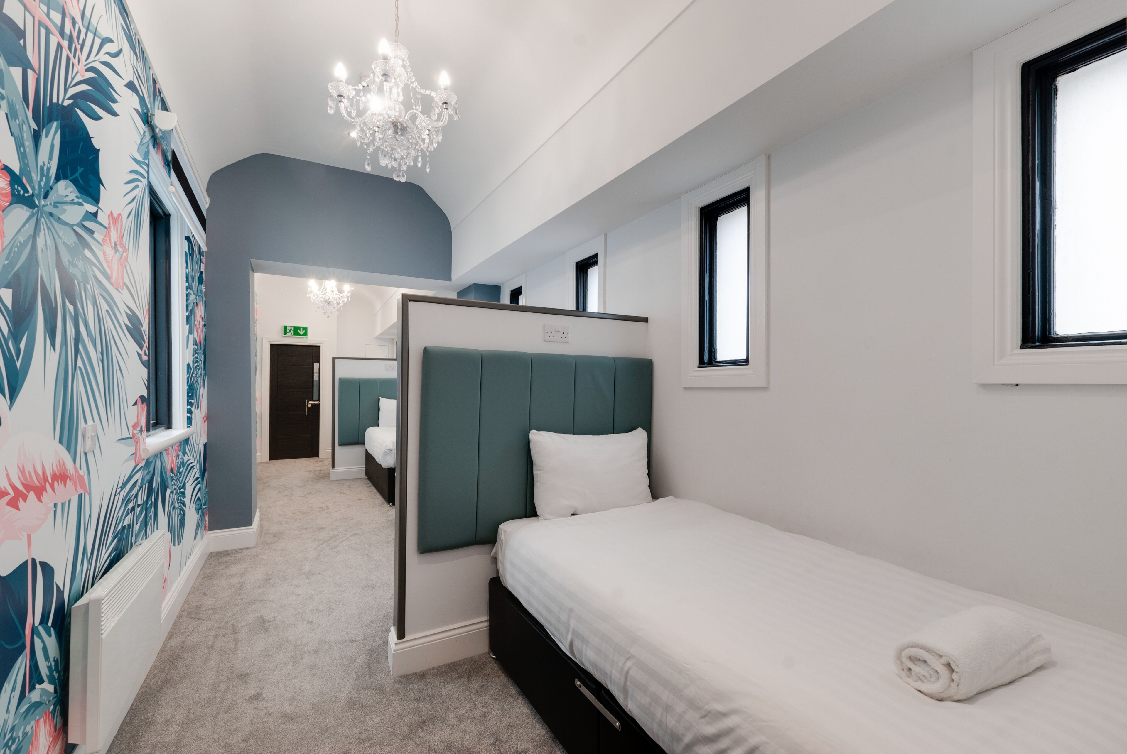 A modern hotel room with a single bed, a green headboard, a white wall, a window, and a chandelier.