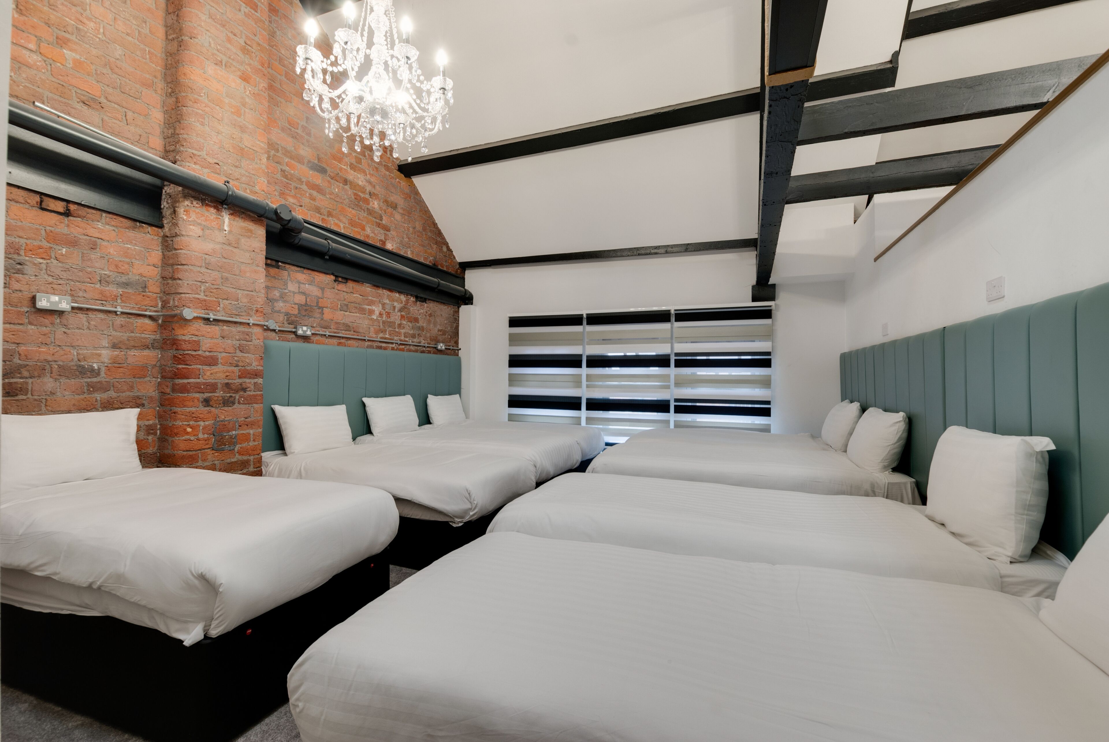 A hotel room with four beds arranged in a row, a brick wall, a modern chandelier, and a large window with blinds.