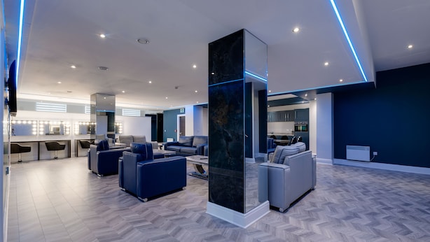 A modern hotel lobby with a herringbone wood floor, a central column with a marble-like texture, and a seating area with sofas and armchairs.