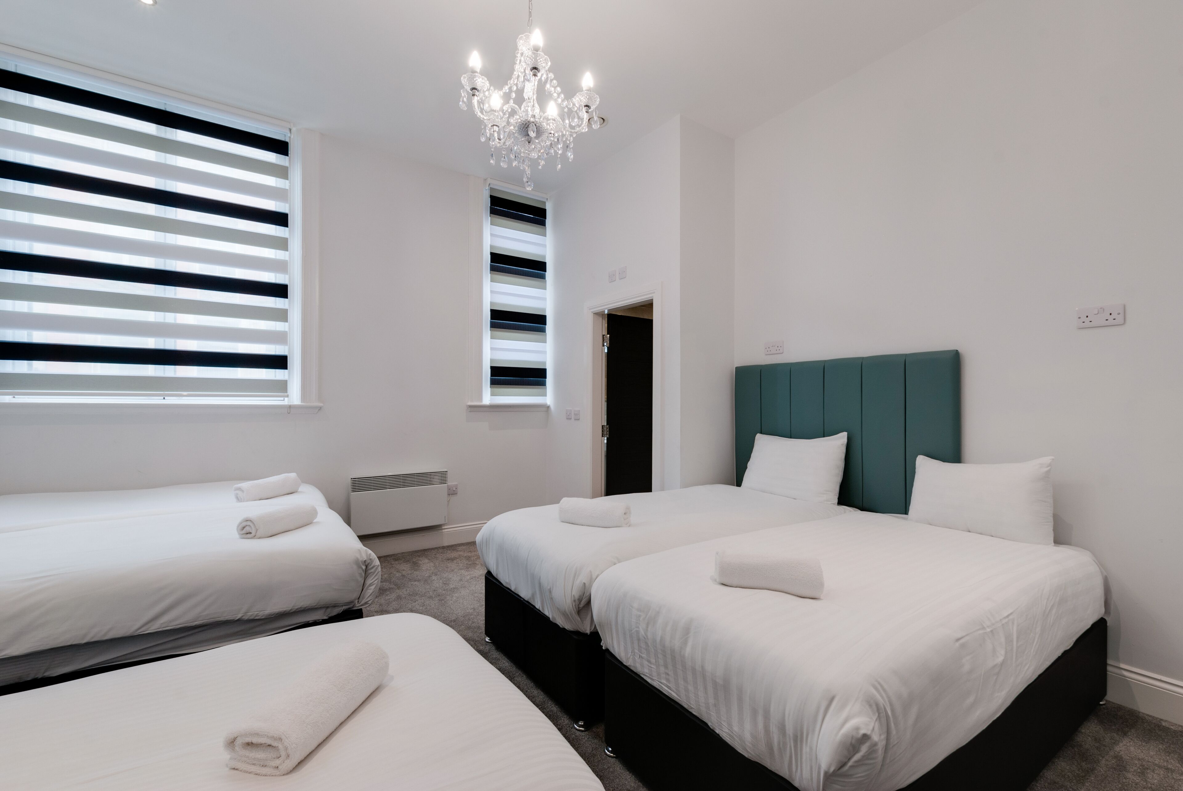 Habitació d'hotel amb dos llits, una làmpada penjant i grans finestres amb persianes.