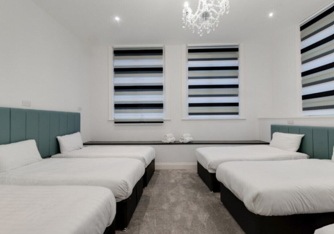Une chambre d’hôtel avec quatre lits simples, une moquette grise, un lustre et deux fenêtres équipées de stores à rayures.