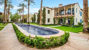A pool surrounded by a manicured lawn and palm trees, with a building featuring balconies and a tiled roof in the background. - Rancho Sol A | 1BR Legacy Villa (La Quinta)
