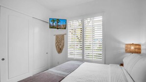 A bedroom with a bed, a wall-mounted TV, a macrame wall hanging, and a window with blinds. - Rancho Sol A | 1BR Legacy Villa (La Quinta)