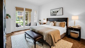 Une chambre à coucher comprenant un lit, un banc, des tables de chevet, un ventilateur de plafond et une fenêtre donnant sur un immeuble.
