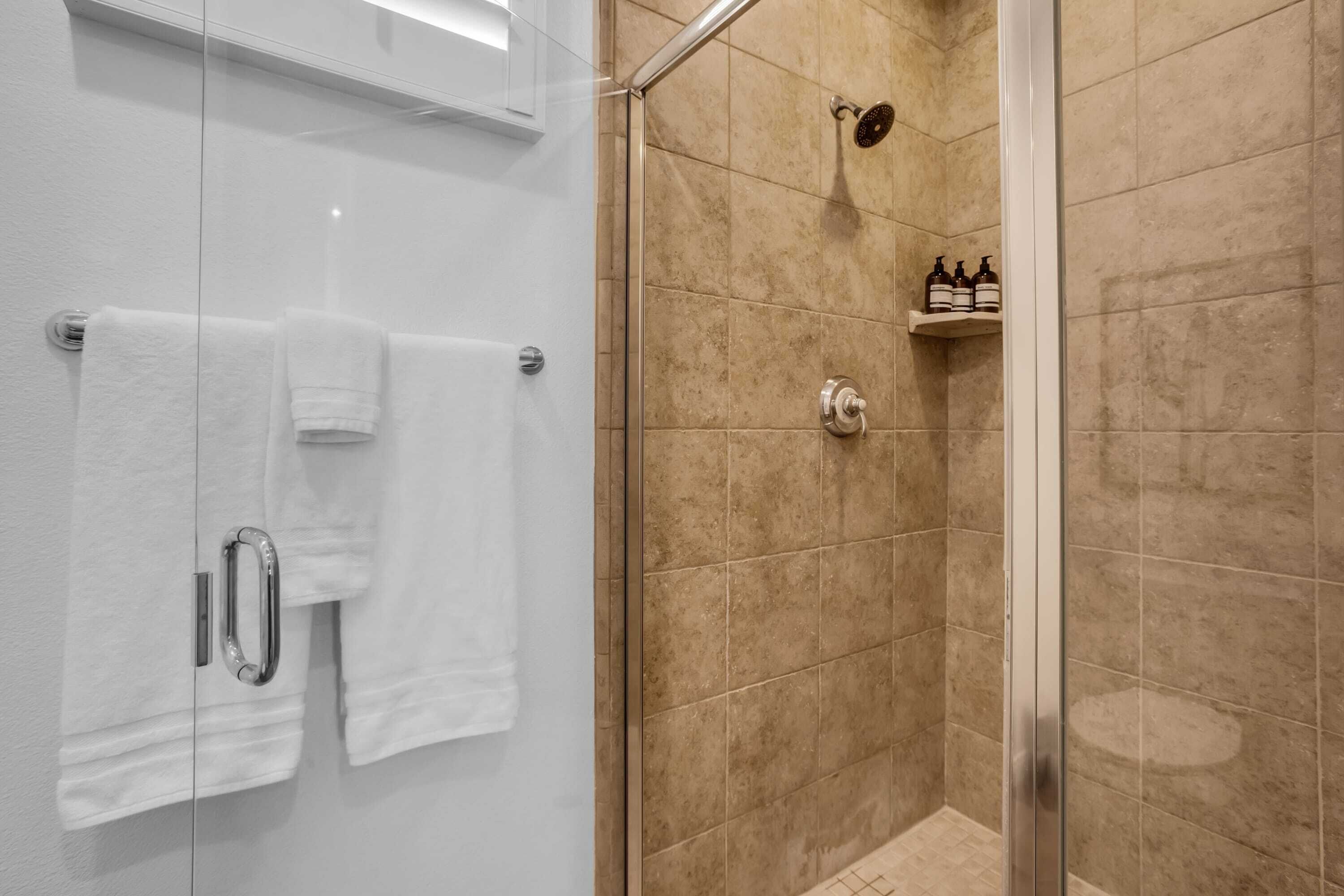 Un espace douche clos par des parois de verre, avec des carreaux de couleur beige, une porte de douche en verre et une étagère contenant des articles de toilette.