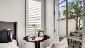 Habitación de hotel con cama, una mesa redonda con lámpara, un cartel enmarcado y vista al exterior.