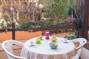 Una mesa redonda con mantel blanco, rodeada de sillas blancas, ubicada al aire libre con vista a follaje y una cerca de madera.