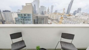 Un balcon avec deux chaises et une petite table, d’où l’on peut admirer un paysage urbain composé d’immeubles modernes et de grues de construction.
