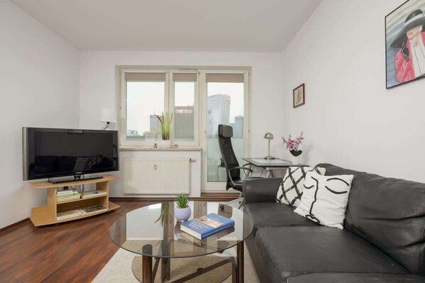 Un soggiorno moderno con una TV a schermo piatto, un tavolino da caffè in vetro, un divano in pelle nera e vista sulla città attraverso ampie finestre.