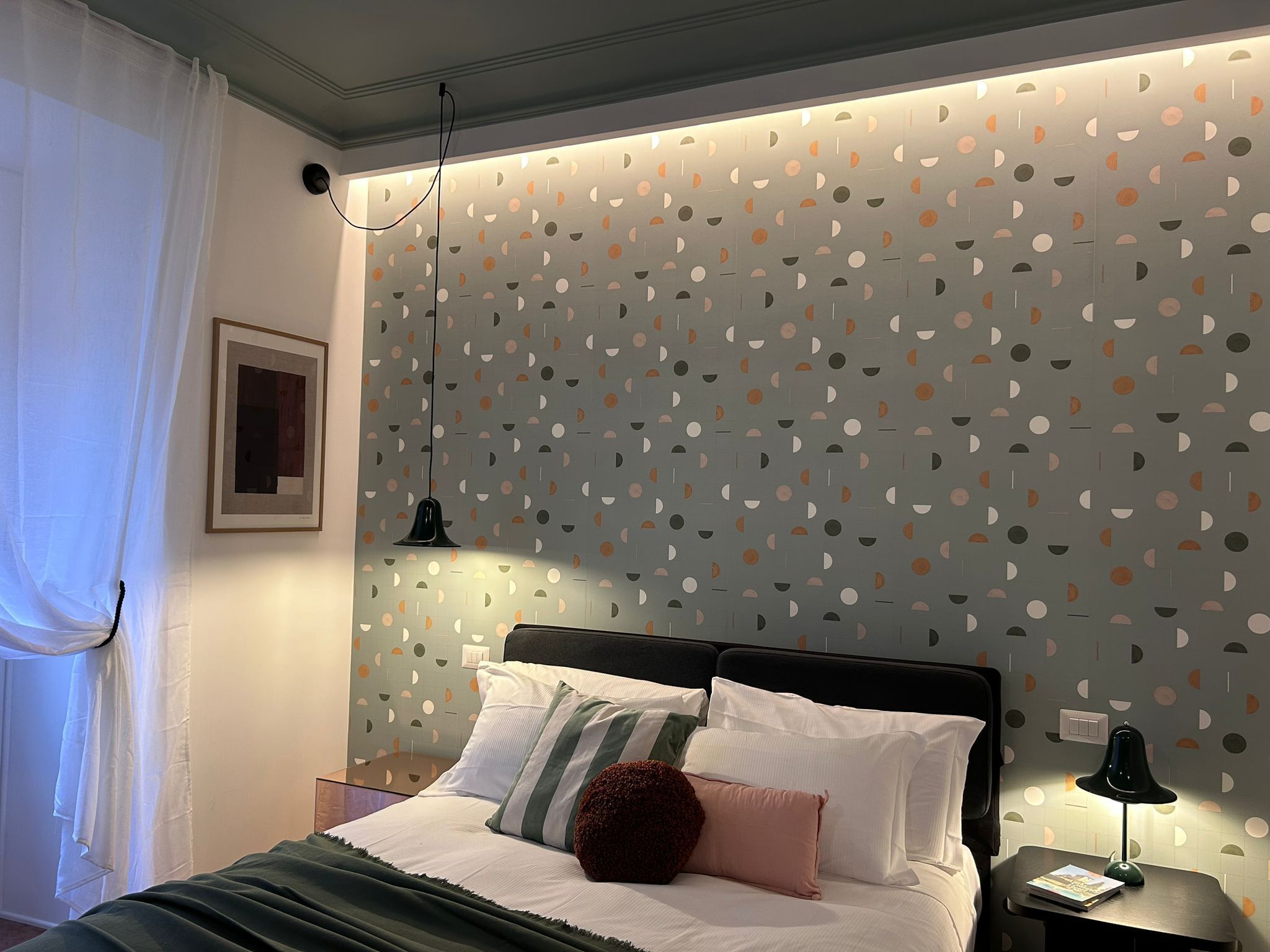 Un lit bien fait, avec des oreillers blancs et à rayures, une couverture verte et un coussin d’accent de couleur rouge foncé. On aperçoit une table de chevet avec une lampe et un livre. Le mur est décoré d’un motif géométrique composé de cercles et de lignes.