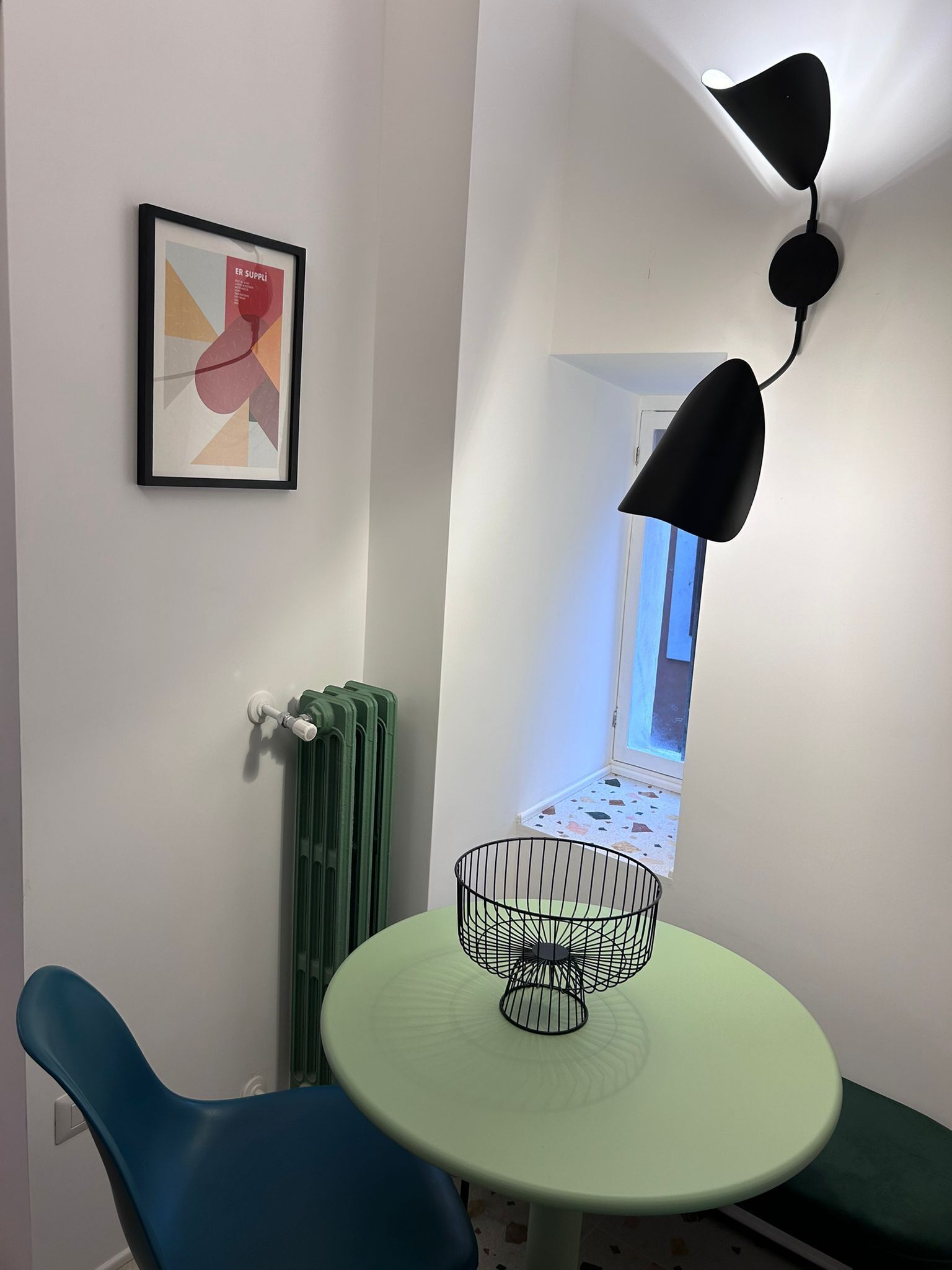 Et lille rum med et grønt bord, en sort stol, en radiator, et billede i ramme på væggen og en sort pendel.
