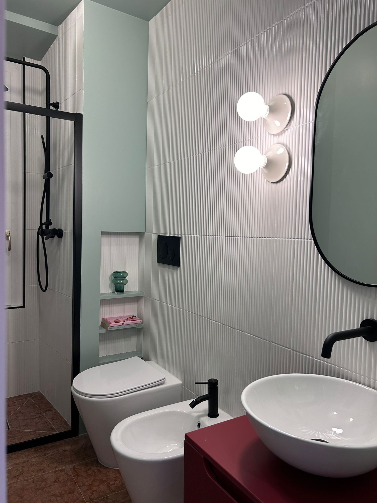 Uma casa de banho com sanita branca, um bidé, uma pia com bancada vermelha e um espelho com luzes.