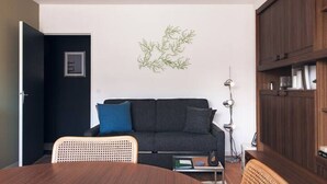 A modern living room with a dark grey sofa, a wooden dining table, and a wall-mounted lamp. - Studio 70, Centre, Proche des Plages (Saint-Palais-sur-Mer)