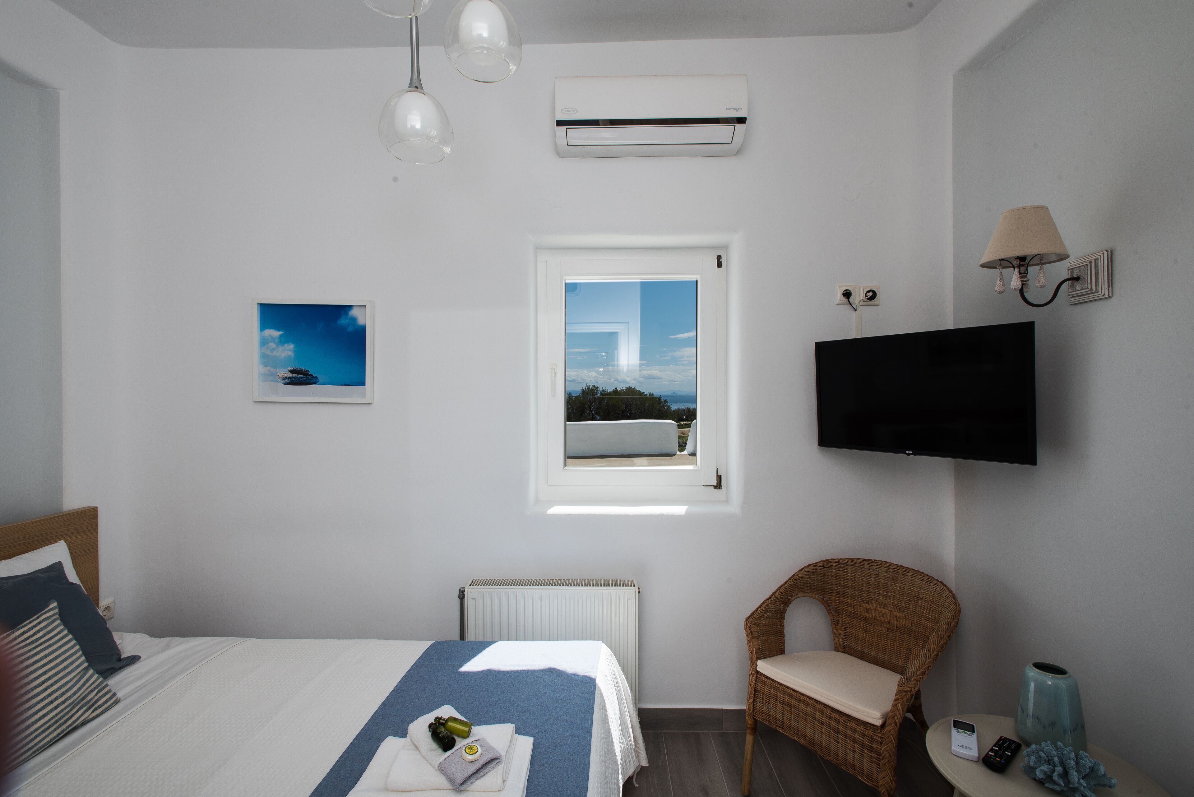 Habitación de hotel con una cama, una silla de mimbre, televisión, aire acondicionado, ventana y un cuadro.