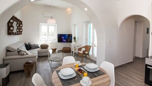 A modern living room with a sofa, dining table, and chairs. There is a television, a window, and a door. - TIFFANYS VILLAS (Mykonos)