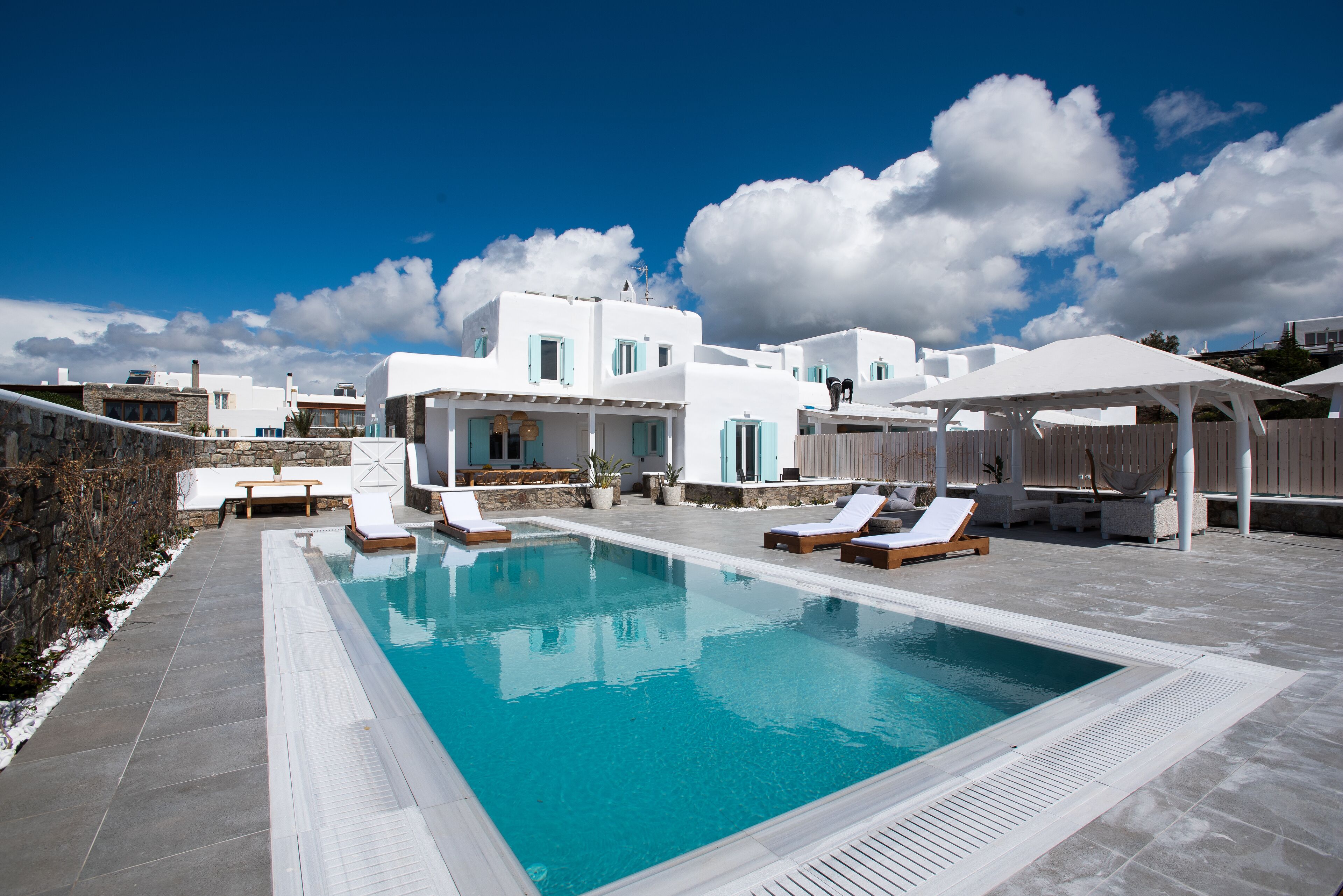 Un área de piscina con sillones de descanso y un edificio con paredes blancas y contraventanas azules.