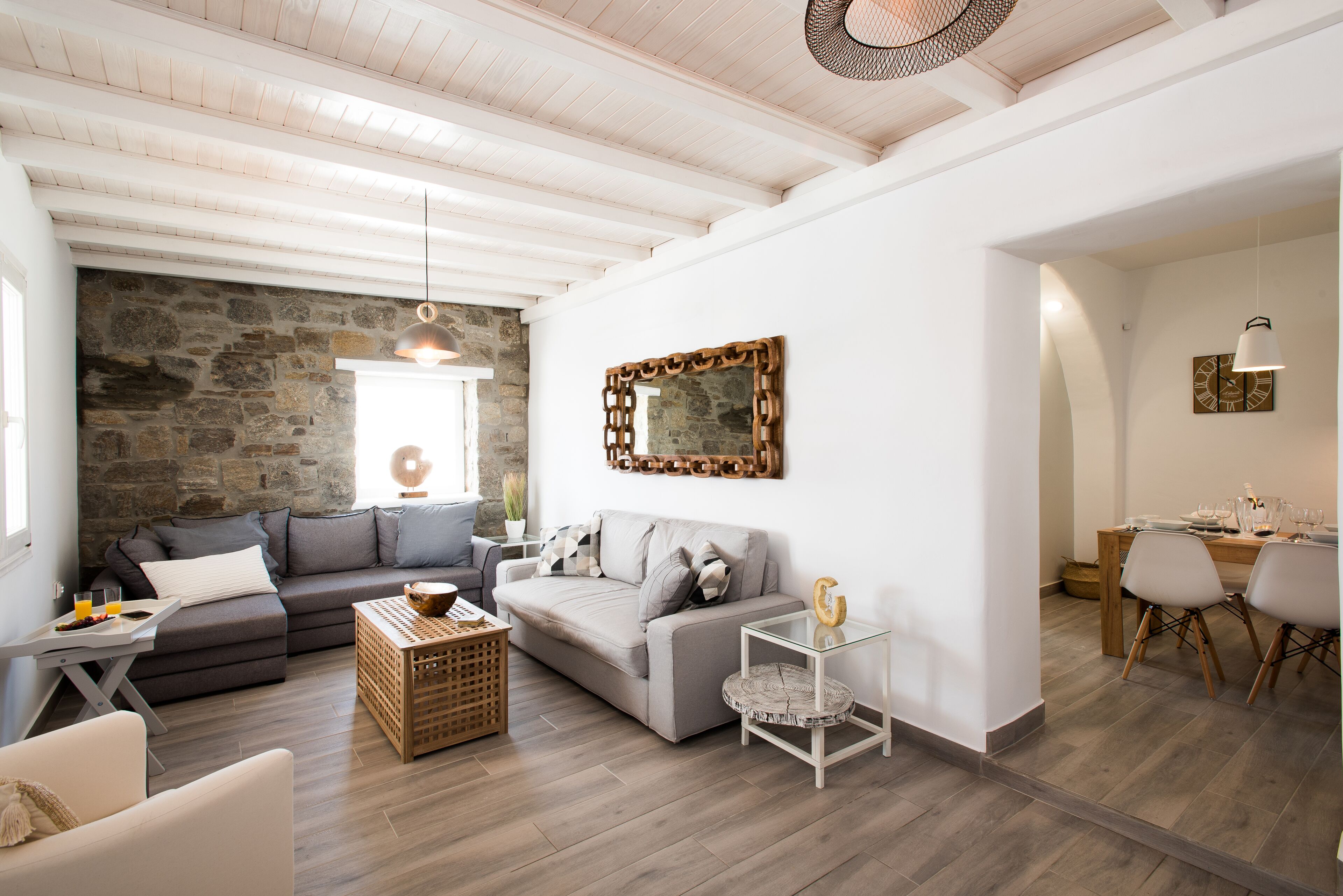 A modern living room with a stone wall, grey sofas, a glass coffee table, and a dining area with wooden chairs and a table.