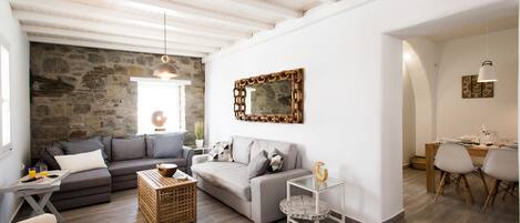A modern living room with a stone wall, grey sofas, a glass coffee table, and a dining area with wooden chairs and a table.
