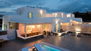A modern white villa with a pool, outdoor seating, and a stone wall. - TIFFANYS VILLAS (Mykonos)