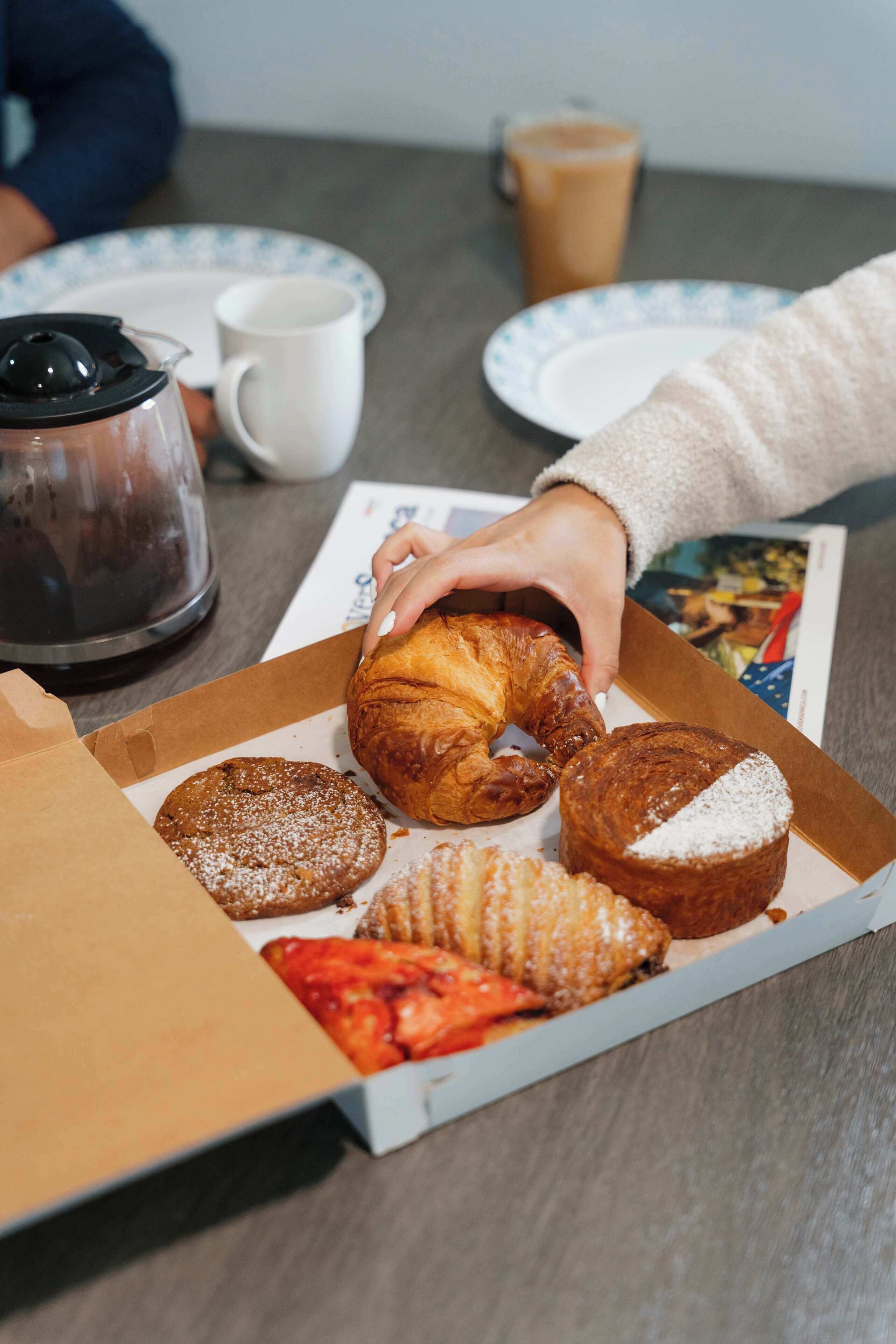 Una caja de pasteles variados sobre una mesa, con una cafetera y una taza al fondo.