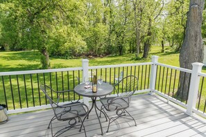 Une terrasse en bois avec une balustrade blanche, deux chaises en métal, une table avec une bouteille et des verres, et une vue sur une pelouse verdoyante et des arbres.