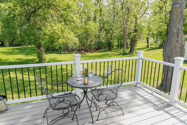 A wooden deck with a white railing, two metal chairs, a table with a bottle and glasses, and a view of a green lawn and trees.