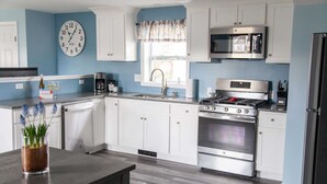 A modern kitchen with white cabinets, stainless steel appliances, a clock on the wall, and a window with a curtain.