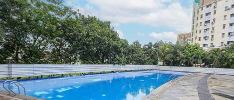 An outdoor swimming pool with a tiled deck, surrounded by trees and a building in the background.