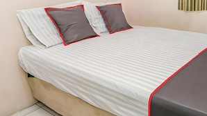 Ein ordentlich bezogenes Bett mit gestreifter Bettwäsche und roter Biese, eine schlichte Wand und ein gestreifter Vorhang.