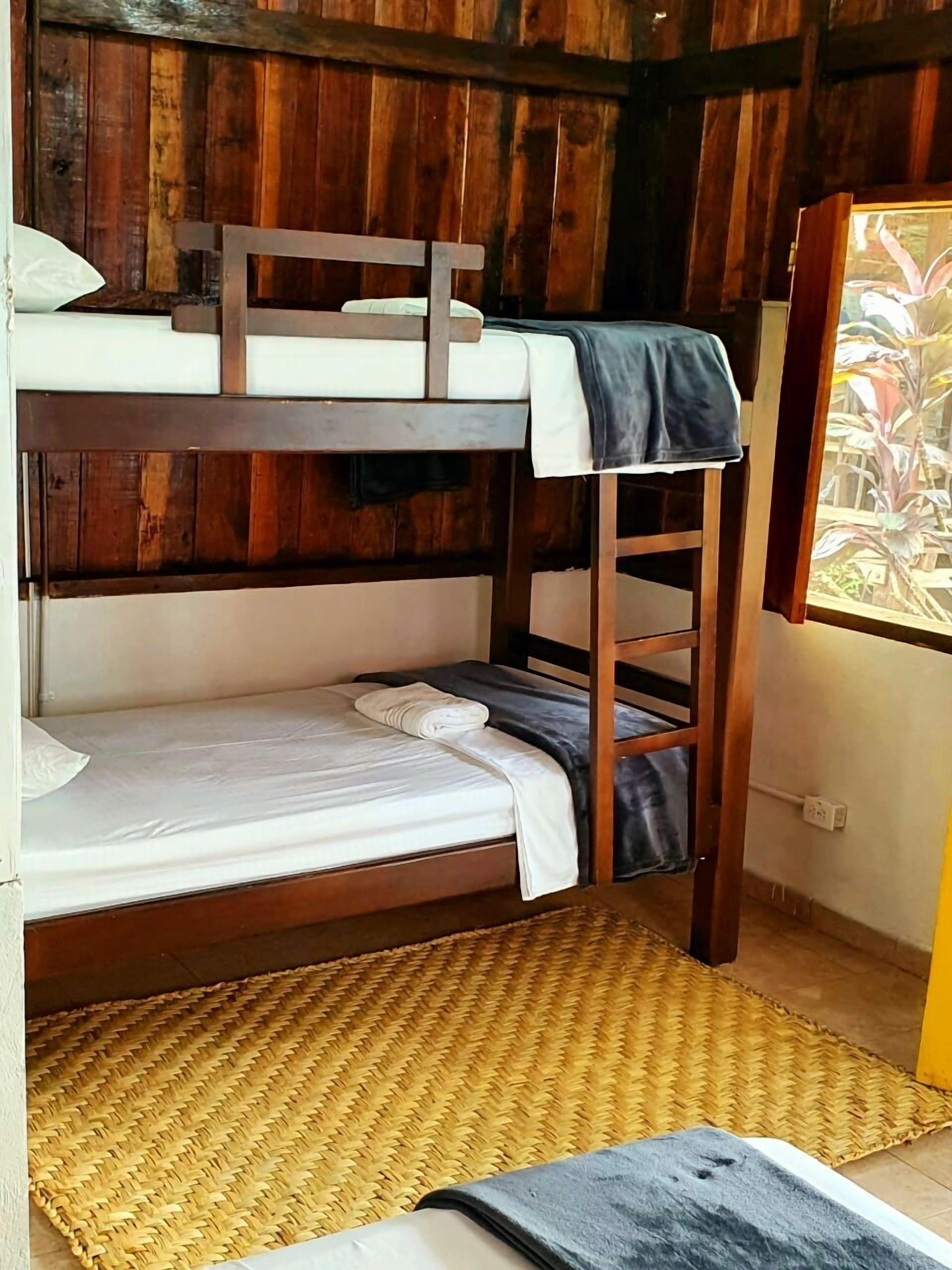 Een stapelbed met een geweven mat op de vloer, houten wanden en een raam met uitzicht.