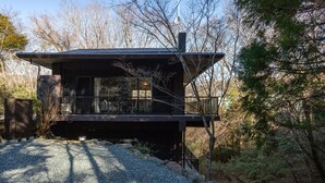 Una casa moderna de dos pisos con un balcón, rodeada por un camino de grava y zona boscosa.