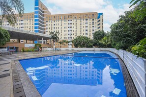 A swimming pool with a clear blue water surface, surrounded by a concrete deck and a low wall. In the background, there is a large multi-story building with numerous windows and balconies. - Collection O 93646 Grand Property By Maria (Tangerang)