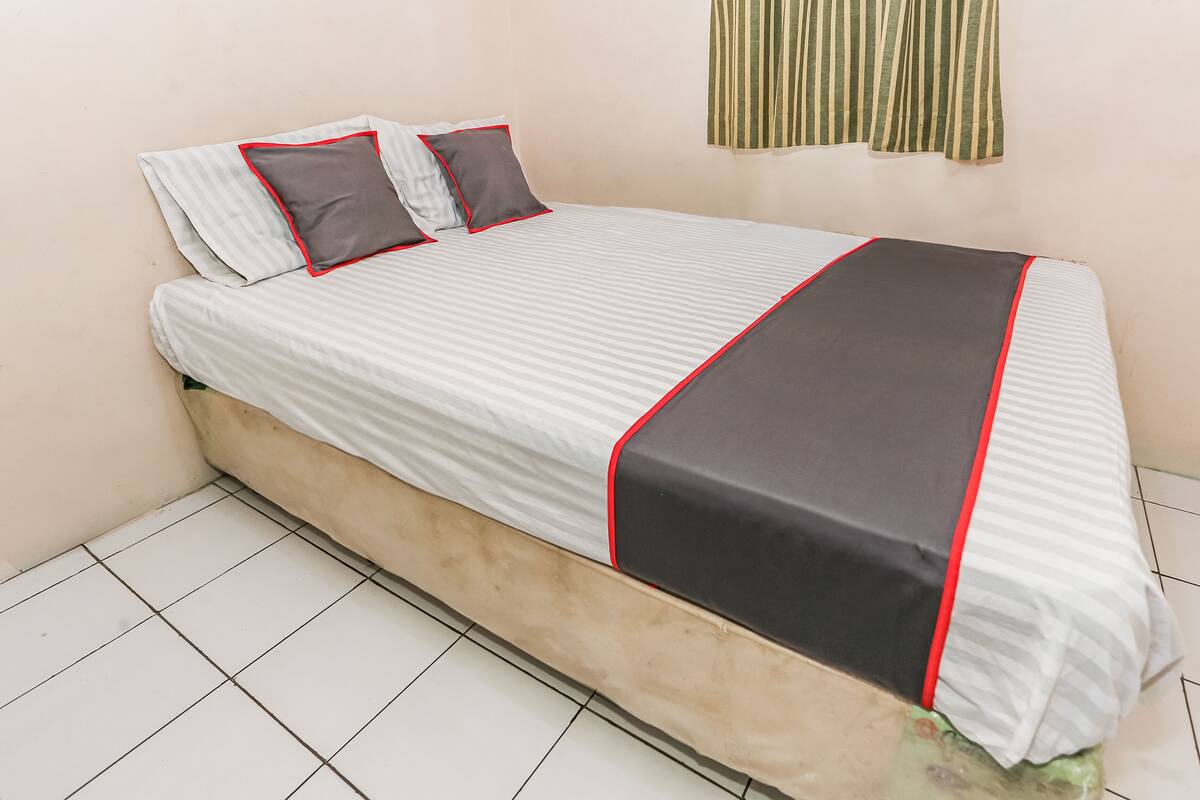 A single bed with a striped bedspread and red trim, two pillows, and a green-striped curtain.