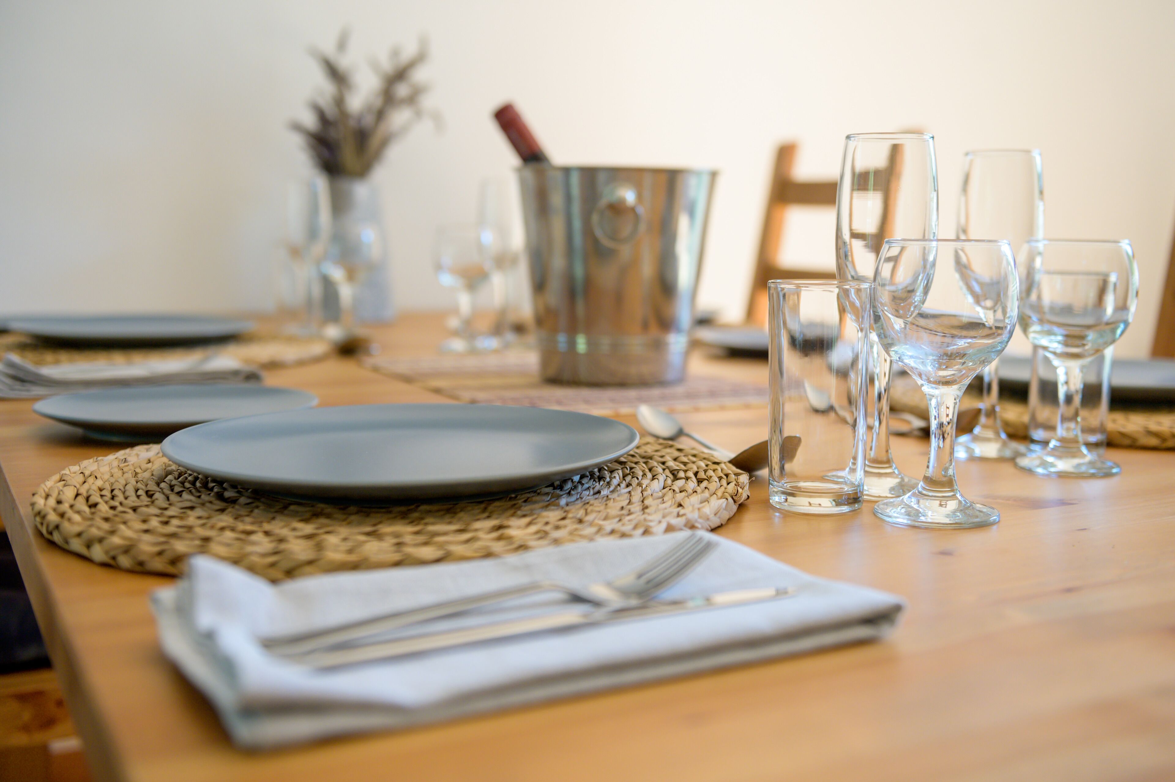 A dining table set with plates, cutlery, and wine glasses, with a metal bucket and a vase in the background.
