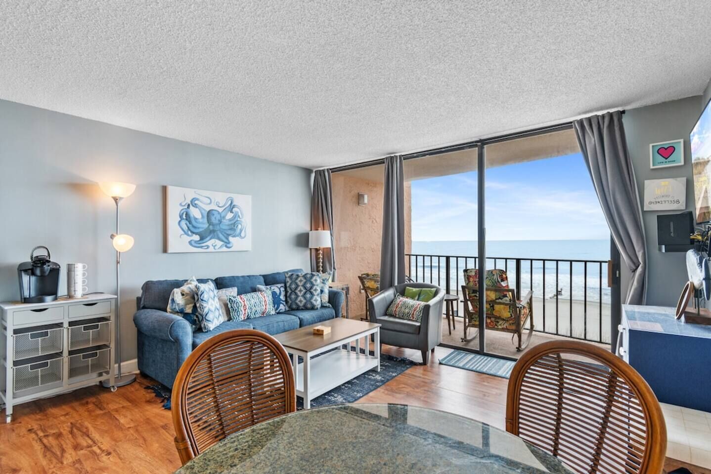 Un salon meublé comprenant un canapé, une table basse et des fauteuils, une table à manger, un coin cuisine et un balcon avec vue sur la mer.