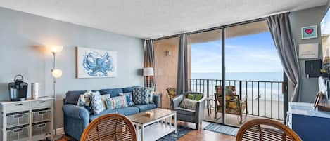 Un salon meublé comprenant un canapé, une table basse et des fauteuils, une table à manger, un coin cuisine et un balcon avec vue sur la mer.