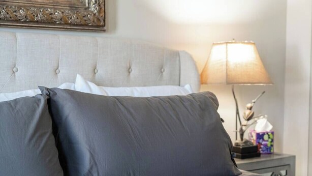 Una cama con ropa de cama gris y un cabecero, una lámpara de noche y una mesita con una caja de pañuelos.