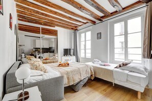 A modern bedroom with a large bed, a smaller bed, a sofa, a TV, and a kitchen in the background.