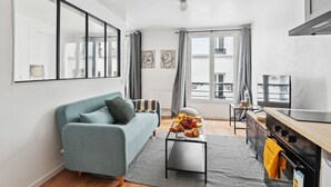 A modern living room with a blue sofa, a coffee table with fruits and pastries, a kitchen with a stove and oven, and large windows with curtains.