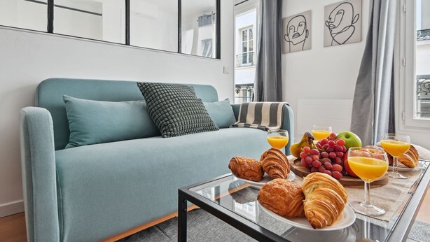 A modern living room with a teal sofa, a glass coffee table with croissants, fruit, and orange juice.