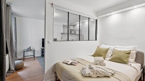 A modern bedroom with a large bed, a bedside table, a window with blinds, and a view of a kitchen area.