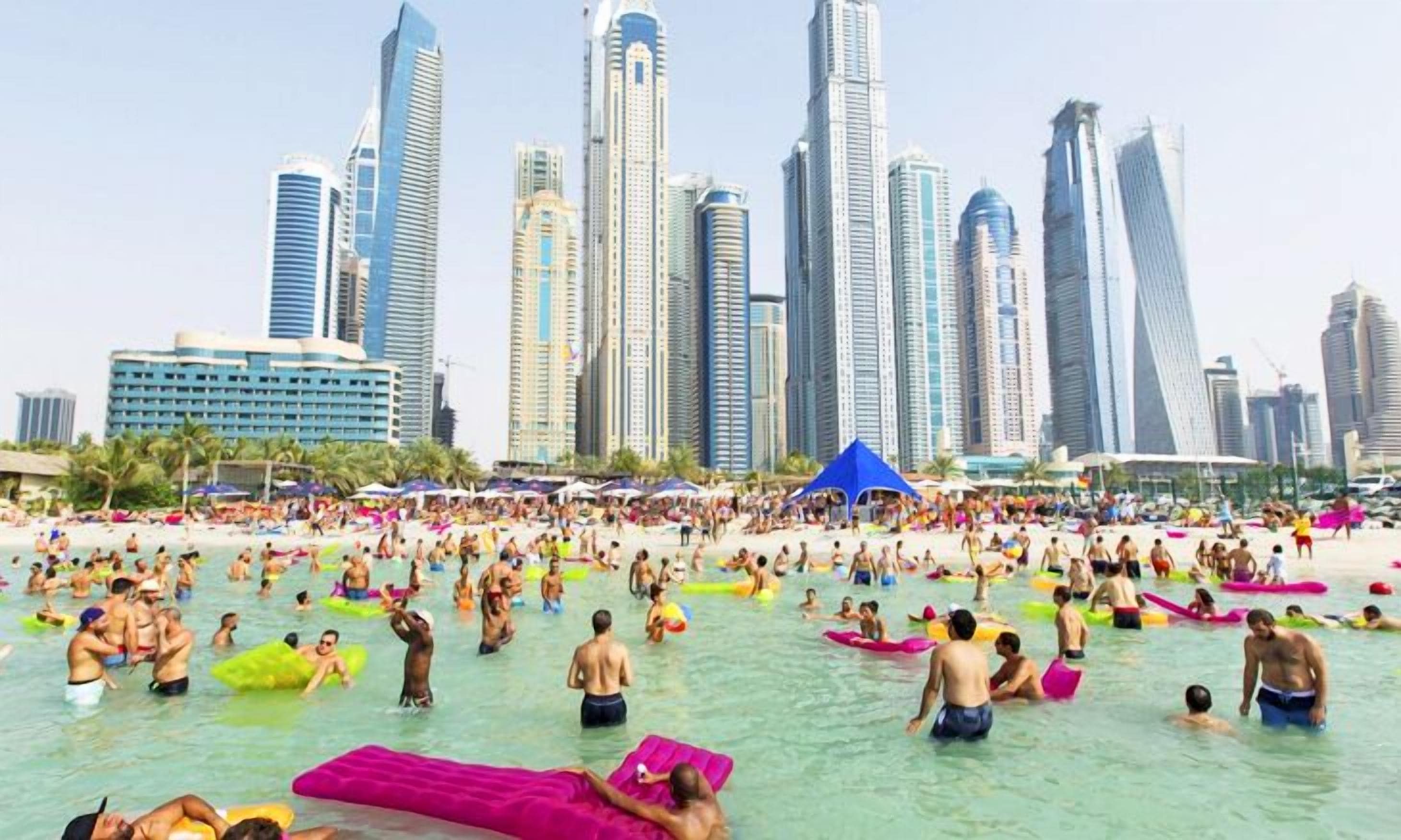 Une plage bondée où l’on voit des gens se baigner et utiliser des bouées, le tout sur fond d’immenses gratte-ciel modernes.