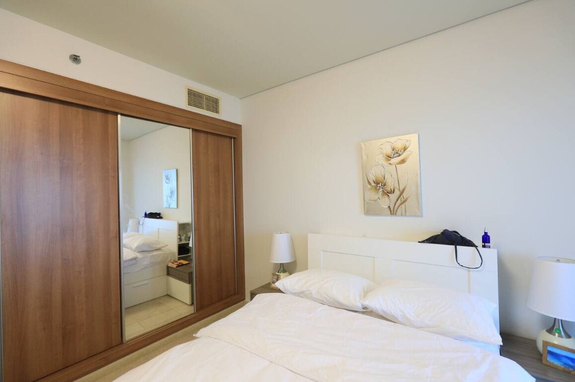 Une chambre d’hôtel dotée d’un grand miroir, d’un lit avec du linge de lit blanc, d’une table de chevet avec une lampe et d’une œuvre d’art accrochée au mur.